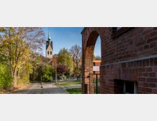 Straßenszene mit einer Kirche im Hintergrund, deren Turm mit einer grünen Spitze versehen ist, umgeben von herbstlich gefärbten Bäumen. Im Vordergrund ein historischer Backsteinbogen, daneben ein gepflasterter Weg, der an Grünflächen und einem Zaun vorbeiführt.