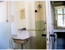 Ein einfacher Waschraum mit einem weißen, wandmontierten Waschbecken, das an einer verblassten Wand mit gelblicher und weißer Farbaufteilung angebracht ist. Im Hintergrund befindet sich ein Fenster mit Holzrahmen, das Tageslicht in den Raum lässt, sowie eine teilweise geöffnete Tür.