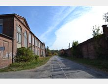 Straße mit Schienen, flankiert von alten Fabrikgebäuden aus rotem Backstein, mit zugemauerten Fenstern und vereinzeltem Bewuchs; im Hintergrund Wälder und ein klarer Himmel.