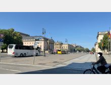 Straßenansicht eines Platzes mit historischen Gebäuden, darunter ein großes, klassizistisches Gebäude mit Säulenfassade. Im Vordergrund fahren ein weißer Reisebus und ein gelber Linienbus, während Radfahrer und Fußgänger den Bereich nutzen.