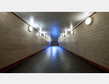 Unterführung mit hell beleuchteten Wänden aus beigefarbenen Kacheln, symmetrisch angeordneten runden Wandlampen und einem Fliesenboden; am Ende führen Stufen nach oben.