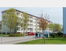 Mehrstöckiges Wohngebäude mit hellem Putz und Balkonen, umgeben von parkenden Autos und mehreren Bäumen auf einer grünen Rasenfläche. Im Vordergrund laufen zwei Personen auf einem Fußweg, und im Hintergrund sind weitere Wohnhäuser und eine ruhige Wohngegend erkennbar.