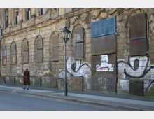 Fassade eines alten, verfallenen Gebäudes mit zugemauerten Fenstern, bröckelndem Putz und Graffiti. Auf dem Gehweg davor läuft eine Person in einem langen Mantel neben einer Straßenlaterne.