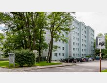 Mehrstöckiges Wohngebäude mit hellgrauer Fassade, umgeben von Bäumen und Grünflächen. Im Vordergrund befinden sich geparkte Autos, Gehwege und Schilder, darunter ein Parkplatzschild.