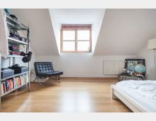 Dachzimmer mit Holzboden, einem Dachfenster mit Holzrahmen, einem weißen Heizkörper darunter und mehreren Möbeln, darunter ein Regal mit Büchern und Kleidung, ein schwarzer Sessel, ein niedriges Bett und ein Nachttisch mit Globus und Lampe.