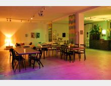 Großer, offener Veranstaltungsraum mit Tischen und Stühlen in der Mitte, einer entspannten Sitzecke links sowie einer Bar im Hintergrund. Der Raum ist durch bunte Beleuchtung und farbenfrohe Dekorationen gestaltet, mit Vorhängen und Kunstwerken an den Wänden.