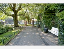 Parkeingang mit gepflastertem Weg, flankiert von großen Bäumen, grünen Rankgittern und Laternen. Im Vordergrund eine weiße Bank und ein Mülleimer, im Hintergrund eine durchbrochene Metallzaunanlage mit öffentlichem Straßenbereich erkennbar.