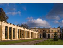 Historisches Gebäude mit einer langen Glasgalerie und klassischen Säulen, flankiert von Statuen in Nischen; im Hintergrund ein großer Torbogen mit dekorativen Elementen. Die Architektur ist von symmetrischem Design, eingebettet in eine grüne Parklandschaft unter einem bewölkten blauen Himmel.