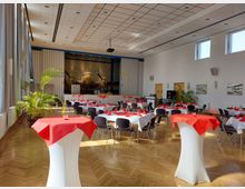 Ein Veranstaltungsraum mit hellen Wänden und großen Fenstern, eingedeckt mit runden Tischen, weißen Tischdecken und roten Servietten. Im Hintergrund ist ein Flugzeugmodell vor einer Bühne und metallischen Vorhängen zu sehen, mit dekorativen Pflanzen am Rand des Raums.