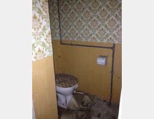 Kleine Toilette mit veralteter Einrichtung. Die Wände sind zur Hälfte mit Holz verkleidet und darüber mit floraler Tapete dekoriert; die Toilette hat einen Deckel mit Steindekor, und Rohre verlaufen an den Wänden.