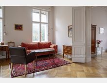 Helles Wohnzimmer mit hohen Decken, großen Sprossenfenstern und Parkettboden. Möbel umfassen ein rotes Sofa, einen Sessel, einen antiken Holztisch auf einem roten Orientteppich und mehrere klassische Holzmöbelstücke; an den Wänden hängen Gemälde.