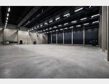 Große, leere Industriehalle mit Betonboden, hohen Decken und freiliegenden Stahlträgern. An der Rückwand befinden sich mehrere schwarze Rolltore und Notausgangstüren, die von Deckenbeleuchtung erhellt werden.