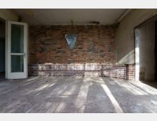 Ein leerer Raum mit einer freiliegenden Ziegelwand, teilweise verkleidet mit dunkleren Kacheln im unteren Bereich. Auf der linken Seite befindet sich eine offene Tür mit Milchglas, während auf der rechten Seite ein Durchgang ohne Tür sichtbar ist; Sonnenlicht wirft Schattenmuster auf den Betonboden.