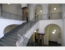 Treppenhaus in einem historischen Gebäude mit verzierten Metallgeländern, runden Marmorsäulen und weißen Wänden. Die Decke ist mit Bögen und Gewölben gestaltet, und mehrere kugelförmige Deckenleuchten hängen von oben.