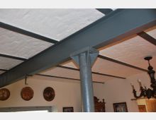 Decke eines Innenraums mit sichtbarer Konstruktion aus dunkelgrauen Stahlträgern und einer weiß verputzten Ziegeldecke, kombiniert mit Dekorelementen wie metallenen Wandtellern und einem Kronleuchter. Im Hintergrund befinden sich Möbel und gerahmte Bilder an den Wänden.