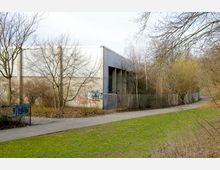 Industriehalle mit grauen Wänden, teils mit Graffiti versehen, in einem halb verwilderten Außenbereich mit Bäumen ohne Laub. Ein Gehweg führt an einem Maschendrahtzaun und einer Grünfläche entlang.