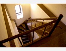 Holztreppe in einem engen Treppenhaus mit weiß gestrichenen Wänden und einem kleinen, bogenförmigen Fenster, das diffuses Licht hereinlässt. Die Treppe ist spiralförmig aufgebaut und verfügt über ein schlichtes hölzernes Geländer.