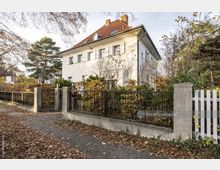 Zweistöckiges Wohnhaus mit hellem Putz und rotem Satteldach, umgeben von einem gepflegten Garten mit Bäumen und Sträuchern. Der Vorgarten ist durch einen Metallzaun mit Steinsäulen begrenzt, und der Gehweg davor ist mit Herbstlaub bedeckt.