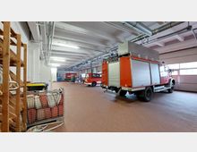 Innenraum einer Feuerwache mit geparkten roten Einsatzfahrzeugen, darunter ein großes Löschfahrzeug und ein kleinerer Transporter. Der Raum hat eine hohe Decke mit Sicht auf Lüftungsrohre, helle Beleuchtung und gefliesten Boden; im Vordergrund sind aufgestapelte Ausrüstungsgegenstände zu sehen.
