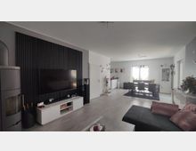 Modernes Wohnzimmer mit Essbereich; links ein großer Fernseher an einer schwarzen Holzpaneelwand, darunter ein weißes Lowboard und ein Kamin. Rechts der Essbereich mit einem Tisch und sechs schwarzen Stühlen vor einem Fenster mit Vorhängen, umgeben von weißen Möbeln und Dekorationen.