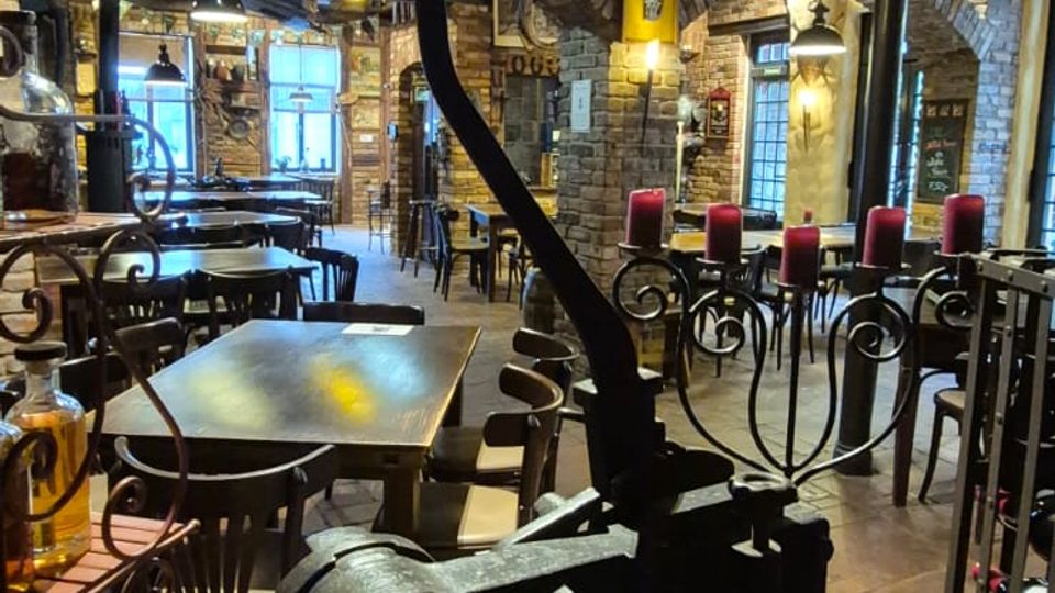 Rustikaler Innenraum eines Restaurants mit Backsteinbögen, sichtbaren Deckenbalken und industriellen Rohren unter der Decke. Im Vordergrund steht eine große schwarze Presse oder Maschine, dahinter Tische und Stühle sowie schmiedeeiserne Kerzenhalter mit roten Kerzen auf Fliesenboden.