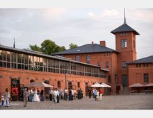 Backstein-Gebäudekomplex mit großen Fenstern im oberen Bereich und einem Turm auf der rechten Seite. Im Innenhof mit Kopfsteinpflaster stehen Personen in kleinen Gruppen, teils unter Sonnenschirmen, und ein Eingang ist mit roten Ballons dekoriert.