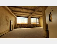 Ein leerer, verlassener Raum mit großflächigen Fenstern, durch die diffuses Licht einfällt. Die Wände sind abgenutzt und teilweise beschädigt, der Boden zeigt Abnutzungsspuren, und eine offene Tür im Vordergrund gibt den Blick in den Raum frei.