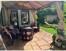 Überdachte Terrasse mit Sitzmöbeln aus dunklem Holz und einem Glastisch auf einem Steinboden. Im Hintergrund ein Garten mit einer Liege auf einer Rasenfläche und einer mit Kletterpflanzen bedeckten Mauer.