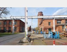 Ehemaliges Industriegelände mit roten Backsteingebäuden, darunter eine große Halle und ein Turm im Hintergrund. Metallkonstruktionen und industrielles Material wie blaue Gestelle und Rohre sind auf einem betonierten Platz verteilt, umgeben von Bäumen und Grünflächen.