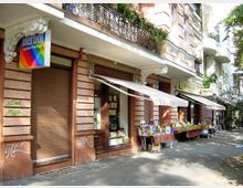 Straßenszene mit einer Altbaufassade, die mit dekorativen Stuckelementen verziert ist; im Erdgeschoss befinden sich Geschäfte, darunter ein Antiquariat mit Büchertischen unter gestreiften Markisen. Daneben ist ein Ladenschild mit der Aufschrift „Herbol Lacke + Farben“ sichtbar.