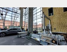 Innenraum eines modernen Gebäudes mit hoher Glasfassade und großen Betonpfeilern. Im Vordergrund stehen ein graues Auto und technische Geräte auf Werkbänken, im Hintergrund ist ein Backsteingebäude durch die Fenster sichtbar.