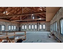 Renovierter Dachraum mit freiliegender Holzbalkenkonstruktion, hellen Wänden und mehreren hohen, spitzbogigen Fenstern, die Tageslicht hereinlassen. Der Raum ist leer und weist Baumaterialien am Rand auf.