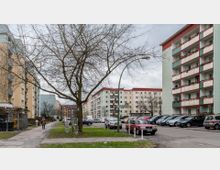 Wohngebiet mit Mehrfamilienhäusern aus mehreren Etagen, teilweise in pastellfarbenen Fassaden. Im Vordergrund steht ein Baum ohne Laub, daneben geparkte Autos und ein Gehweg, auf dem eine Person zu Fuß geht.