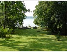 Große, offene Rasenfläche in einem Park, umgeben von hohen, dicht belaubten Bäumen, mit Blick auf ein Gewässer im Hintergrund. Ein Aussichtspunkt mit Brüstung befindet sich am Rand des Wassers.