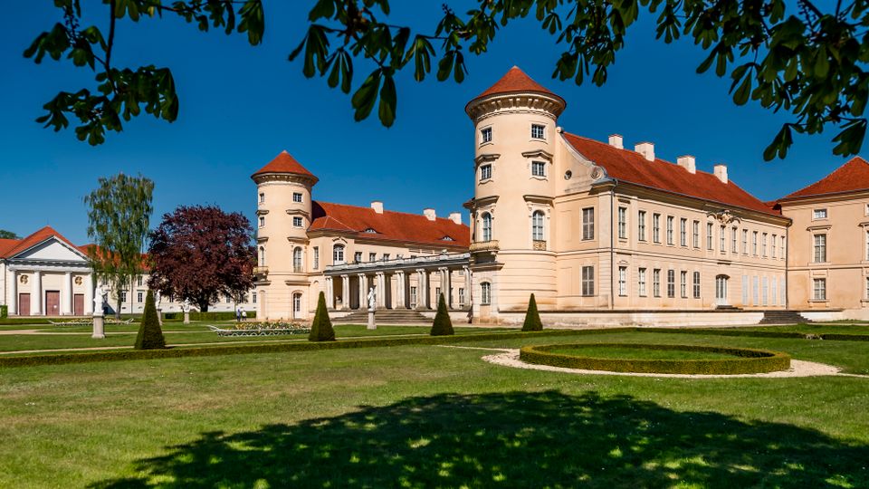 Wide view of a pale stucco palace with red-tiled roofs and round corner towers, fronted by a columned entrance. Formal landscaped lawns with trimmed hedges and small conical shrubs surround the building, with a smaller classical pavilion to the left under a clear blue sky.