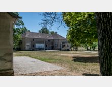 Ein einfaches, zweistöckiges Gebäude mit Putzfassade und Satteldach steht in einer ländlichen Umgebung, umgeben von Bäumen und einer grasbewachsenen Fläche. Im Vordergrund befinden sich ein gepflasterter Weg und einige unbefestigte Flächen.