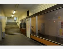 Treppenaufgang in einem U-Bahn- oder Unterführungstunnel mit weißen Kachelwänden, Deckenbeleuchtung und gläsernen Schaufenstern auf der rechten Seite. Graffiti und Plakate sind an den gläsernen sowie gemauerten Flächen sichtbar.