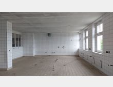 Ein leerer Raum mit weißen, gefliesten Wänden und beigem Fliesenboden, vermutlich eine ehemalige Küche oder ein Arbeitsbereich. An den Wänden befinden sich freiliegende Anschlüsse und Kabel, sowie mehrere Fenster auf der rechten Seite, die natürliches Licht hereinlassen.