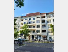 Mehrstöckiges Wohnhaus mit weißer Fassade und rotem Ziegeldach, umgeben von Bäumen. Im Erdgeschoss befinden sich Geschäfte, darunter ein Second-Hand-Laden; Fahrräder sind vor dem Gebäude abgestellt.