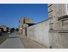 Straßenszene in einem städtischen Wohngebiet mit einfacher Bebauung; rechts im Bild verläuft eine verputzte Mauer, daneben ein Bürgersteig und eine Straße. Im Hintergrund sind Wohnhäuser und eine Kirchturmspitze unter klarem blauen Himmel zu sehen.