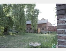 Hof mit einer Backsteinscheune, die ein Ziegeldach hat und Holztüren sowie kleine Fenster zeigt. Im Vordergrund ist eine grüne Wiese mit einer alten Weide, einer Holzbank, einem Liegestuhl und einem runden Holzelement zu sehen.