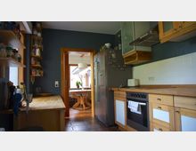 Moderne Küche mit Holzschränken, einer Arbeitsplatte und Fliesenrückwand. Im Hintergrund führt eine offene Tür zum Esszimmer mit Holztisch und Tageslicht.