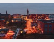 Städtische Szene bei Dämmerung mit roten Backsteingebäuden und einem modernen Bau mit leuchtenden Fenstern. Im Hintergrund ist ein Kirchturm mit spitzem Dach zu sehen, während im Vordergrund Bahngleise und mit Graffiti versehene Fassaden dominieren.