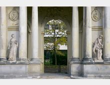 Steingebäude mit großen Säulen und zwei Statuen, die links und rechts in Nischen stehen. Im Hintergrund ein Torbogen, durch den ein Baum sowie ein modernes Gebäude sichtbar sind.