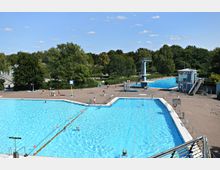 Freibad mit mehreren großen Außenpools, umgeben von einer gepflasterten Fläche und Bäumen im Hintergrund. Sichtbar sind ein Sprungturm, ein kleines Gebäude und mehrere Badegäste, sowie klare blaue Himmel.