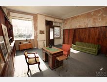 Ein einfacher, rustikal eingerichteter Raum mit abgenutzten Wänden, großen Fenstern und Teppichboden. Im Raum stehen ein Holzschreibtisch mit einem Telefon, verschiedene Stühle, eine grüne Sofacouch, ein Raumteiler aus Holz und ein niedriger Schrank.