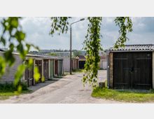 Eine Reihenanlage mit alten Garagen aus Holz und Stein, teilweise mit unterschiedlichen Farbtönen an den Toren, auf einem unbefestigten Weg. Im Vordergrund hängen Zweige eines Baumes, im Hintergrund sind moderne Wohngebäude und bewölkter Himmel zu sehen.