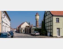 Straßenszene in einem Dorf mit Kopfsteinpflaster, geschlossenen Wohnhäusern im Fachwerkstil und einem gelben Wasserturm mit rotem Kegeldach im Hintergrund. Geparkte Autos stehen entlang der Straße.
