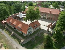 Luftaufnahme eines historischen Gebäudekomplexes mit Fachwerkhaus und rotem Ziegeldach, umgeben von alten Steinmauern. Im Hintergrund befindet sich ein größeres Backsteingebäude mit Torbogen, die gesamte Anlage ist von Grünflächen und Bäumen umgeben.