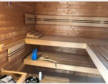 Innenansicht einer traditionellen Sauna aus Holz mit Holzbänken auf mehreren Ebenen. Im Vordergrund sind eine Steinschale für Aufgüsse, ein blauer Wasserkrug und eine hölzerne Kelle sichtbar.